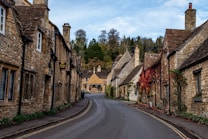 A picturesque village street lined with charming stone cottages, some adorned with climbing plants and colorful foliage. The road gently curves, leading the eye towards more cottages and trees in the background under a partly cloudy sky.