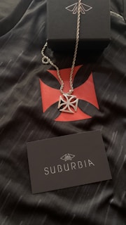 A silver necklace with a geometric pendant is placed on a black fabric featuring a red cross-like design. A black box with a white design is positioned above the necklace. A black card with the word 'SUBURBIA' in white letters and a logo is also visible.