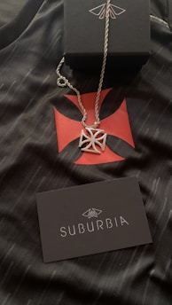A silver necklace with a geometric pendant is placed on a black fabric featuring a red cross-like design. A black box with a white design is positioned above the necklace. A black card with the word 'SUBURBIA' in white letters and a logo is also visible.