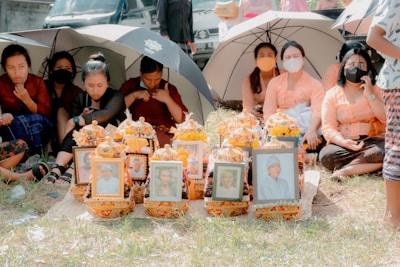A group of people, some wearing masks, sit on the grass under umbrellas. In front of them are floral arrangements and framed photographs, suggesting a memorial or ceremonial gathering.
