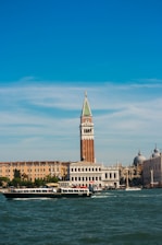 A scenic view of Venetian architecture featuring a prominent bell tower with a green spire against a clear blue sky. Below, a canal filled with boats, including a prominent black and white boat at the forefront. Historical buildings, including a domed structure, line the water's edge.