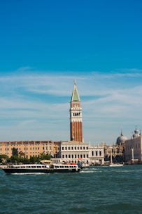 A scenic view of Venetian architecture featuring a prominent bell tower with a green spire against a clear blue sky. Below, a canal filled with boats, including a prominent black and white boat at the forefront. Historical buildings, including a domed structure, line the water's edge.
