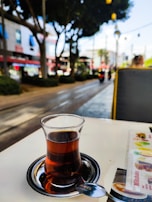 A small glass of tea sits on a table with a metallic saucer and a spoon beside it. The background features a street scene with trees, buildings, and blurred figures, suggesting a lively outdoor setting.