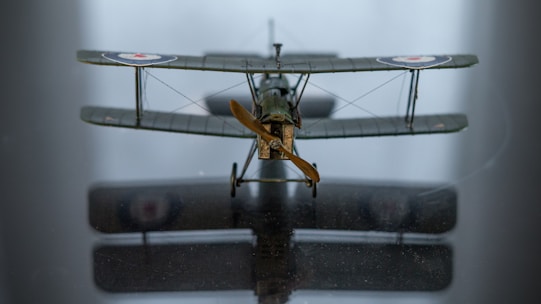 A model biplane is centered on a reflective surface, showcasing its front view. It features two main wings, a detailed propeller, and roundel markings on the wings. The reflection on the surface adds to the intricacy of the image.