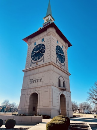 A large clock tower with a pointed green roof, featuring two expansive clock faces with Roman numerals. The structure is adorned with elegant architectural details and the word 'Berne' is prominently displayed on its facade. The scene is set against a clear blue sky, and a few bare trees and neatly trimmed bushes are visible in the vicinity.