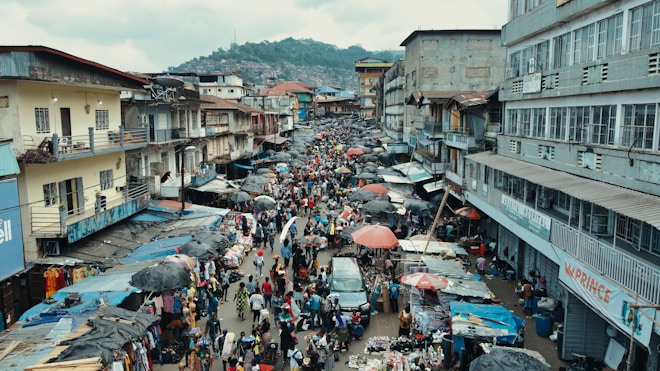 A bustling outdoor market scene with numerous people walking between rows of stalls on a lively street. The market is flanked by multi-story buildings, with vendors selling various goods under umbrellas and makeshift awnings. The area is crowded with vibrant colors from clothing, umbrellas, and merchandise.