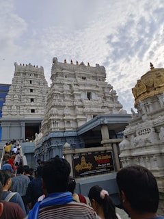 A crowd of people is ascending towards a large, white temple structure characterized by its intricate architectural details. The temple has multiple ornate towers and is adorned with detailed carvings and a golden dome. The sky above is partly cloudy, adding to the scene's atmospheric feel.