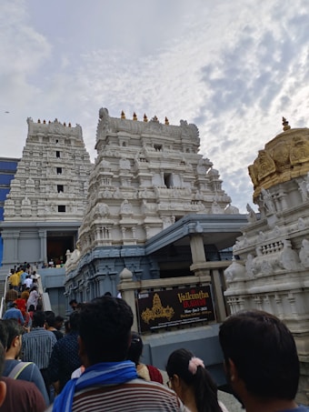 A crowd of people is ascending towards a large, white temple structure characterized by its intricate architectural details. The temple has multiple ornate towers and is adorned with detailed carvings and a golden dome. The sky above is partly cloudy, adding to the scene's atmospheric feel.