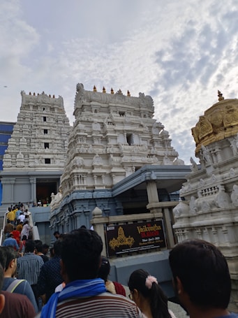 A crowd of people is ascending towards a large, white temple structure characterized by its intricate architectural details. The temple has multiple ornate towers and is adorned with detailed carvings and a golden dome. The sky above is partly cloudy, adding to the scene's atmospheric feel.