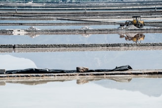 Expansive salt evaporation ponds with a series of rectangular pools reflecting the sky, some with heaps of salt piled up. A yellow industrial vehicle, possibly a backhoe, is present on the right side, adding an industrial element to the scene.