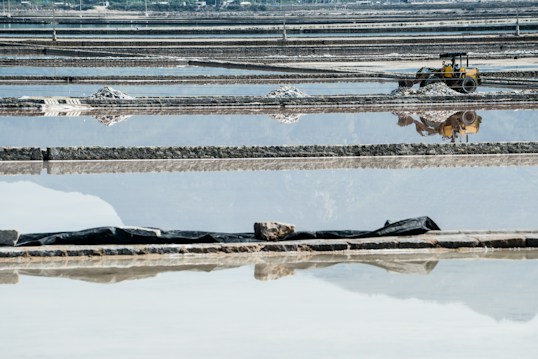 Expansive salt evaporation ponds with a series of rectangular pools reflecting the sky, some with heaps of salt piled up. A yellow industrial vehicle, possibly a backhoe, is present on the right side, adding an industrial element to the scene.