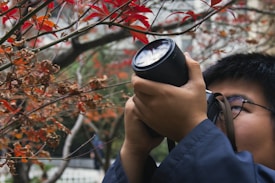A person is holding a camera up to their eye, seemingly taking a photo of or through a branch with red leaves. The scene suggests a focus on nature photography with the background softly blurred.