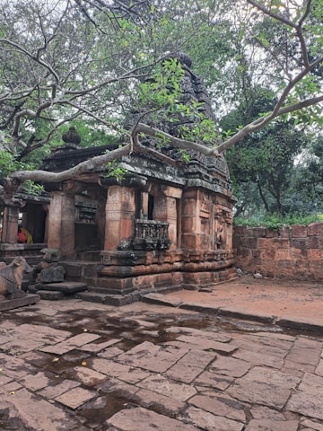 A serene, ancient stone structure surrounded by lush green trees. The temple is adorned with intricate carvings and has a weathered appearance, indicating its historical significance. The stone path in front is slightly wet, suggesting recent rain. The setting feels tranquil and secluded.