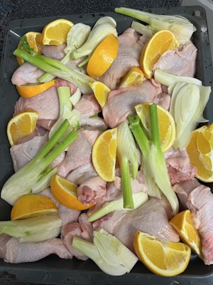 Raw chicken pieces are arranged on a baking tray, surrounded by cut fennel stalks and orange wedges. The colors of the ingredients are fresh and vibrant, with the oranges and fennel adding a contrasting brightness to the pink chicken.