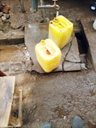 A water tap is mounted on a concrete platform outdoors, with water running into a yellow plastic jerry can. Another jerry can is placed on the ground nearby, and a small wooden stool is visible in the corner. The area is wet with patches of mud and dirt.