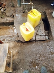 A water tap is mounted on a concrete platform outdoors, with water running into a yellow plastic jerry can. Another jerry can is placed on the ground nearby, and a small wooden stool is visible in the corner. The area is wet with patches of mud and dirt.
