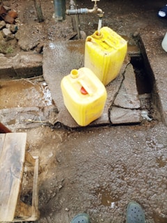 A water tap is mounted on a concrete platform outdoors, with water running into a yellow plastic jerry can. Another jerry can is placed on the ground nearby, and a small wooden stool is visible in the corner. The area is wet with patches of mud and dirt.