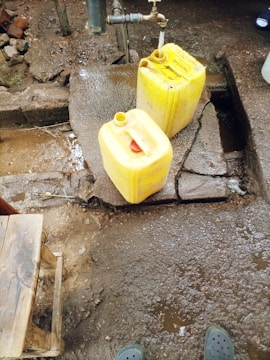 A water tap is mounted on a concrete platform outdoors, with water running into a yellow plastic jerry can. Another jerry can is placed on the ground nearby, and a small wooden stool is visible in the corner. The area is wet with patches of mud and dirt.