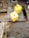 A water tap is mounted on a concrete platform outdoors, with water running into a yellow plastic jerry can. Another jerry can is placed on the ground nearby, and a small wooden stool is visible in the corner. The area is wet with patches of mud and dirt.
