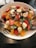 A vibrant fruit salad in a white bowl, featuring a colorful mix of fresh ingredients such as banana slices, strawberries, orange segments, blueberries, and almond slices atop a bed of green leafy vegetables. A silver fork and spoon rest on the sides of the bowl on a dark wooden table.