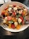 A vibrant fruit salad in a white bowl, featuring a colorful mix of fresh ingredients such as banana slices, strawberries, orange segments, blueberries, and almond slices atop a bed of green leafy vegetables. A silver fork and spoon rest on the sides of the bowl on a dark wooden table.
