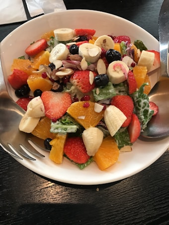 A vibrant fruit salad in a white bowl, featuring a colorful mix of fresh ingredients such as banana slices, strawberries, orange segments, blueberries, and almond slices atop a bed of green leafy vegetables. A silver fork and spoon rest on the sides of the bowl on a dark wooden table.