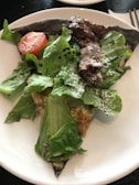 A plate with a slice of pizza topped with fresh green lettuce, a cherry tomato, grilled meat, and sprinkled Parmesan cheese.