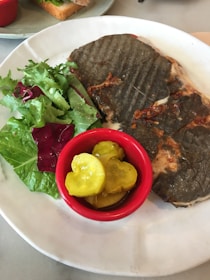 A grilled sandwich with a dark crust is served on a white plate alongside a fresh green salad and a small red cup of pickles.