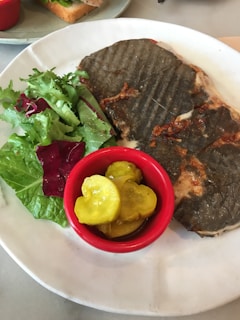 A grilled sandwich with a dark crust is served on a white plate alongside a fresh green salad and a small red cup of pickles.