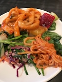 A plate of assorted Korean dishes featuring spicy rice cakes, seasoned noodles, fresh greens, and sliced vegetables. The food is vibrantly colored with orange, red, green, and purple elements.