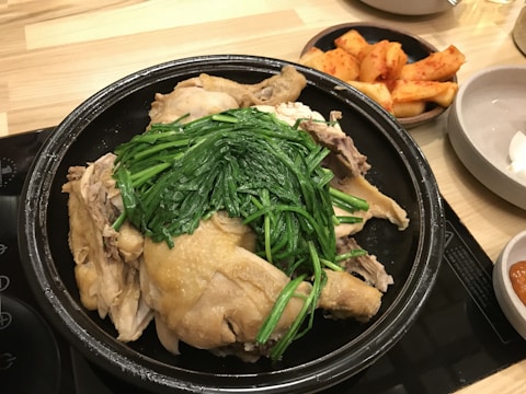 A cooked whole chicken is served in a black pot with fresh green vegetables layered on top. On the side, there is a dish of pickled radish kimchi.