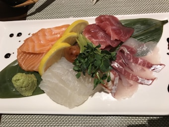 A beautifully arranged platter of sashimi, featuring various slices of raw fish including salmon, tuna, and possibly snapper. The plate is garnished with fresh lemon slices, greens, and wasabi, all laid atop a decorative leaf. Soy sauce is drizzled around the edges of the plate.