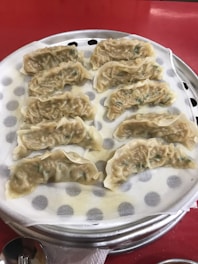 A metal steaming tray containing several dumplings placed on wax paper with small circular holes. The dumplings have a slightly translucent appearance and appear to be filled with a greenish vegetable mixture. The background is a red table surface, and there is a spoon positioned near the tray.