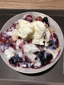 A bowl filled with a dessert containing shaved ice topped with two scoops of vanilla ice cream, mixed berries, and a drizzle of sauce. The dish is garnished with granola on a dark tray.