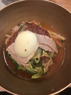 A bowl of food featuring sliced beef, a boiled egg, thinly sliced cucumbers, bean sprouts, and noodles, all in a rich broth. The topping includes a red chili paste, adding a vibrant color contrast.