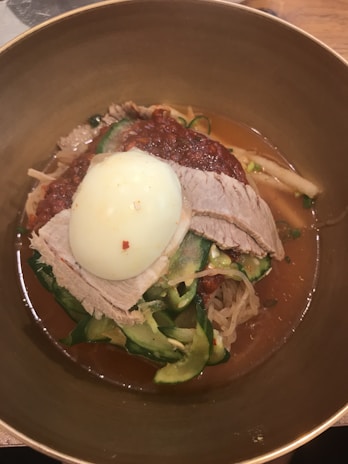 A bowl of food featuring sliced beef, a boiled egg, thinly sliced cucumbers, bean sprouts, and noodles, all in a rich broth. The topping includes a red chili paste, adding a vibrant color contrast.