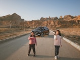 Two children are standing on a road in a desolate area with rocky hills in the background. Behind them, there is a black car parked on the road. The children appear to be holding small objects and are dressed in casual clothing.