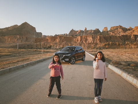 Two children are standing on a road in a desolate area with rocky hills in the background. Behind them, there is a black car parked on the road. The children appear to be holding small objects and are dressed in casual clothing.