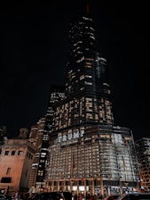 A tall skyscraper is prominent against a dark night sky, with bright windows and a large illuminated sign displaying the word 'TRUMP'. Surrounding the building are other smaller structures and various architectural details, with traffic and people visible at the street level.