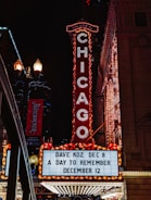 A bright marquee sign against a nighttime urban backdrop, featuring the word CHICAGO in illuminated letters. The venue advertises upcoming shows with artists such as Dave Koz and an event called A Day to Remember. Surrounding elements include ornate architectural details and streetlights, contributing to a lively city atmosphere.