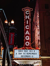 A bright marquee sign against a nighttime urban backdrop, featuring the word CHICAGO in illuminated letters. The venue advertises upcoming shows with artists such as Dave Koz and an event called A Day to Remember. Surrounding elements include ornate architectural details and streetlights, contributing to a lively city atmosphere.