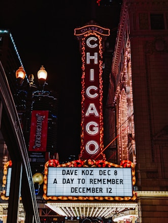 A bright marquee sign against a nighttime urban backdrop, featuring the word CHICAGO in illuminated letters. The venue advertises upcoming shows with artists such as Dave Koz and an event called A Day to Remember. Surrounding elements include ornate architectural details and streetlights, contributing to a lively city atmosphere.