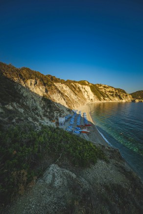 A picturesque coastal scene with a gently sloping beach lined with sun loungers and umbrellas. Steep cliffs rise to the left, covered with patches of grass and bushes, while the sky is a deep, clear blue. The water near the shore is calm, displaying shades of green and blue.