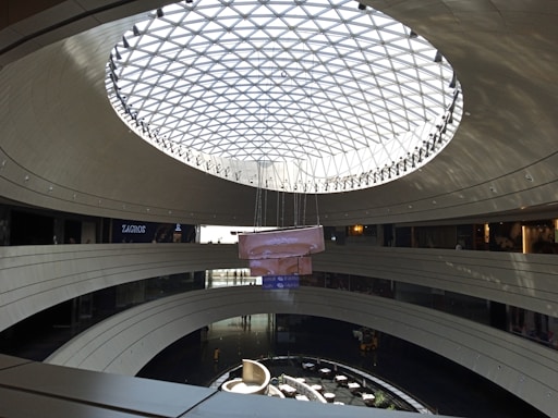 A modern interior with a large, circular atrium, the top of which is covered by a geometric glass dome allowing natural light to illuminate the space. Multiple levels curve around the open center, with a seating area and tables arranged below. The architecture features smooth, gray surfaces and sleek lines.