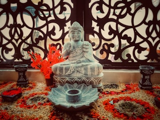 An intricately carved Buddha statue sits in front of a decorative, ornate panel. Bright red flowers are arranged around the statue, which is placed on a woven mat with various floral designs.
