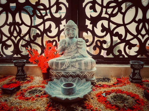 An intricately carved Buddha statue sits in front of a decorative, ornate panel. Bright red flowers are arranged around the statue, which is placed on a woven mat with various floral designs.