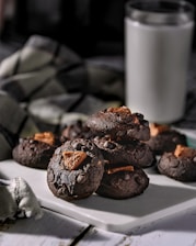 A stack of dark chocolate cookies with chunks of chocolate embedded in them sits on a white cutting board. In the background, there is a blurred glass of milk and a green checkered cloth.