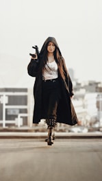 A person walks confidently in an urban setting, wearing a long black coat with a hood and carrying a firearm. The outfit includes a white shirt, black pants, and tall boots with buckles.