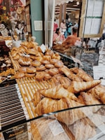 A bakery display is filled with an assortment of pastries, including croissants and cinnamon rolls, all arranged neatly on wooden shelves. Reflection seen on the glass shows people sitting and enjoying their food in a cozy setting, with warm lighting and decorative elements.