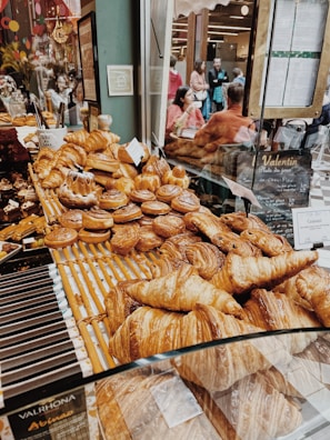 A bakery display is filled with an assortment of pastries, including croissants and cinnamon rolls, all arranged neatly on wooden shelves. Reflection seen on the glass shows people sitting and enjoying their food in a cozy setting, with warm lighting and decorative elements.